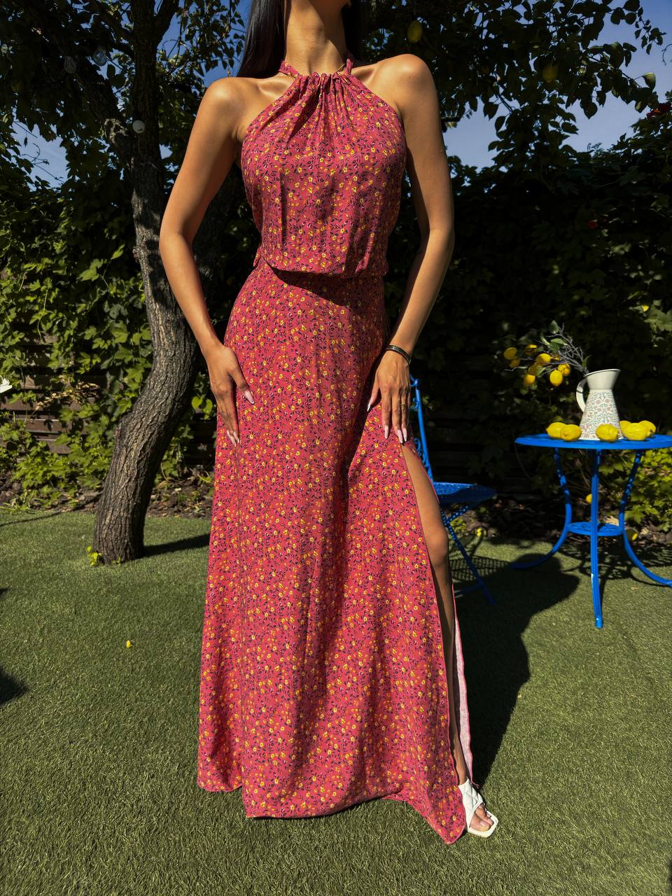 Woman wearing a red floral dress standing outdoors with greenery and a blue chair in the background.