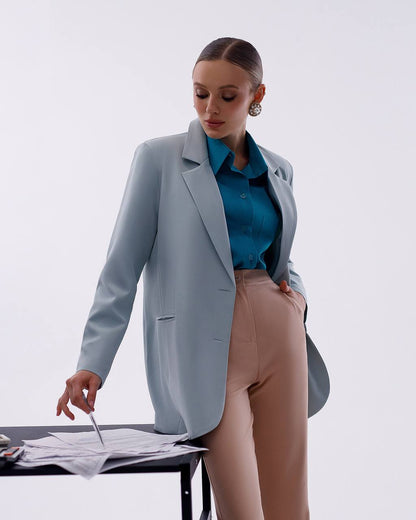 A woman models a light blue blazer over a teal button-up shirt and beige trousers, standing next to a desk with papers on a plain light gray studio background.
