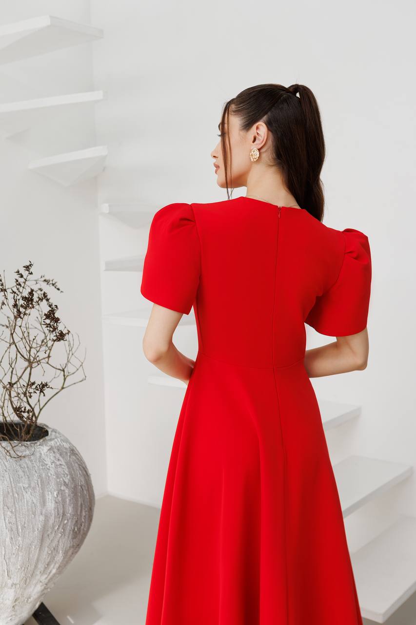 Woman in a red dress standing in a minimalistic room with a white wall and a decorative plant.