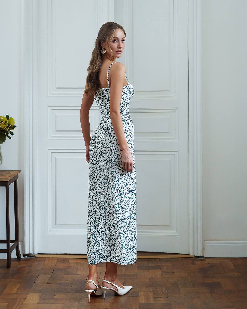 Woman in a floral dress standing in a room with wooden flooring and white walls.