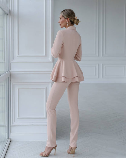 Woman wearing a light pink peplum jacket and pants in a room with white paneled walls.