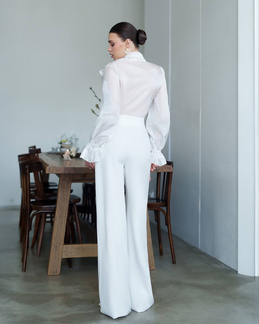 Woman in a white outfit standing in a minimalistic room with a wooden table and chairs.
