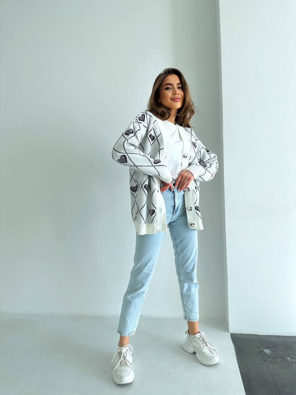 Woman wearing a white patterned jacket, light blue jeans, and white sneakers in a minimalistic setting.