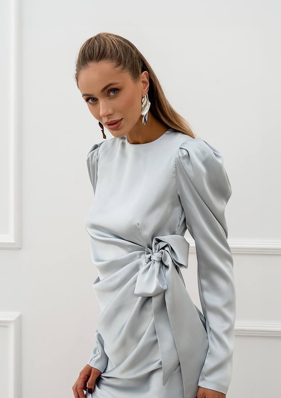 Woman wearing a light blue dress with long sleeves and a tie detail against a white wall.