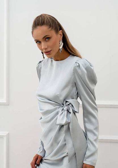 Woman wearing a light blue dress with long sleeves and a tie detail against a white wall.