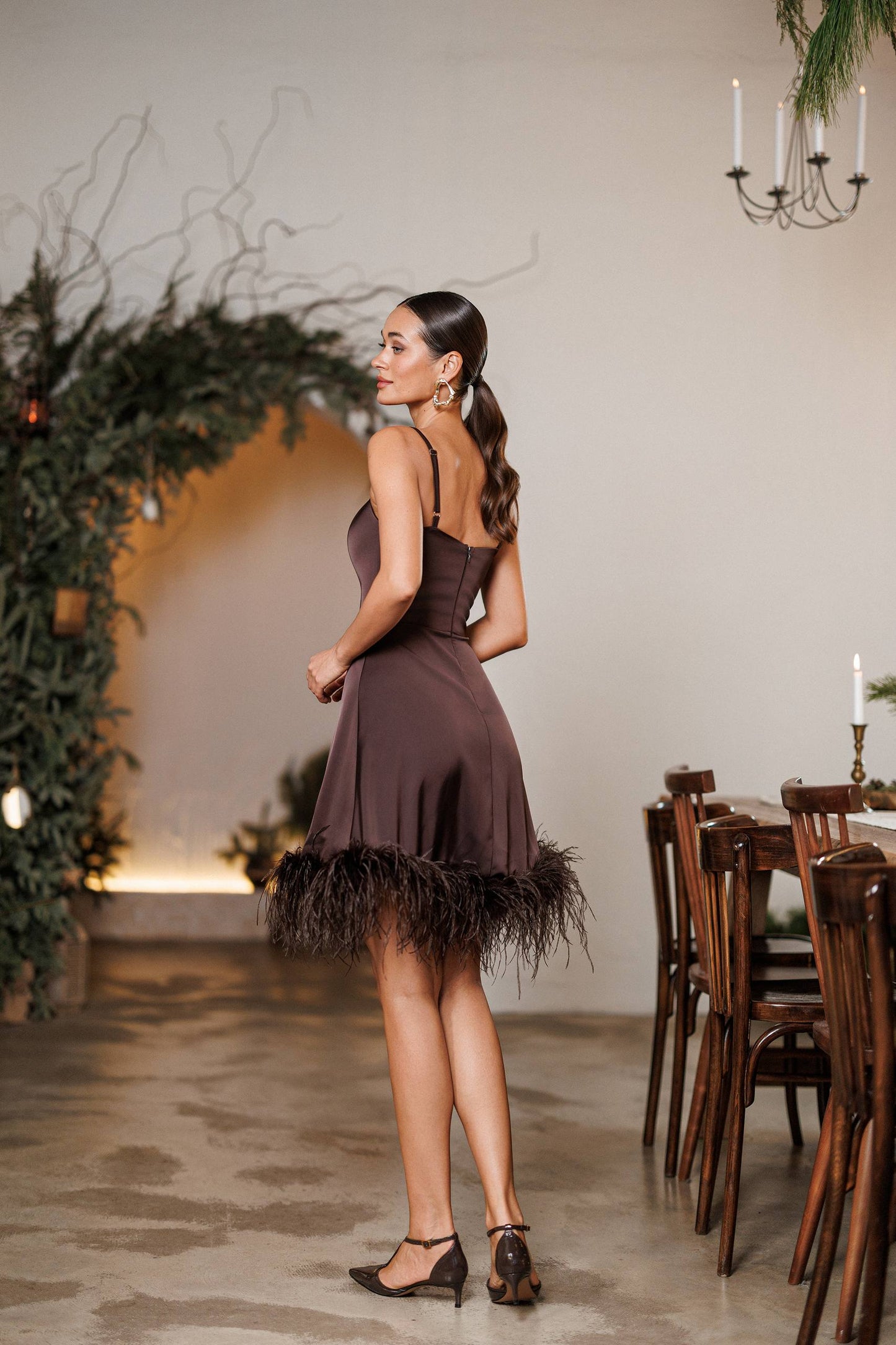 Woman in a brown dress with feathered hem standing in an elegant indoor setting.