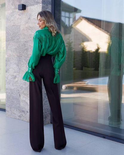 Woman wearing a green blouse and black pants standing in front of a glass door.