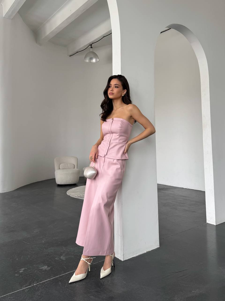 Woman in a pink strapless top and high-waisted pants standing against a white wall.