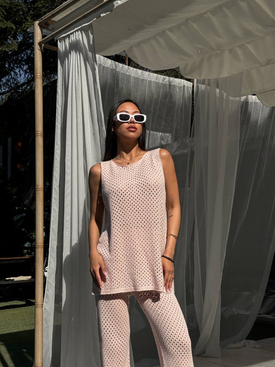 Woman in a light pink sleeveless top and pants standing under a white canopy with sheer curtains.