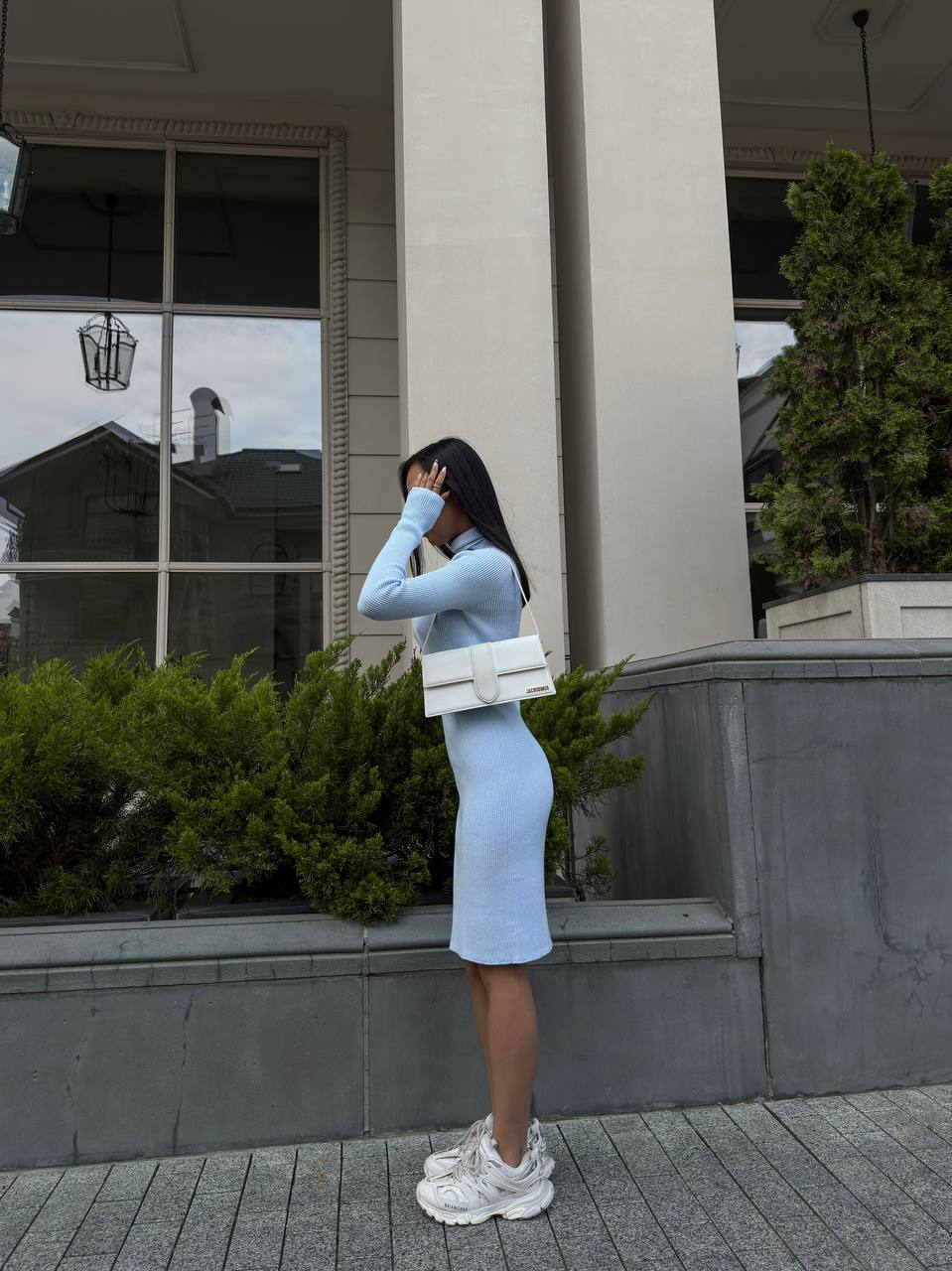 Woman in a light blue dress standing outside a building with greenery.