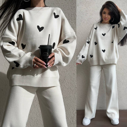 Woman wearing a white sweater with black heart patterns and white pants, holding a black coffee cup.