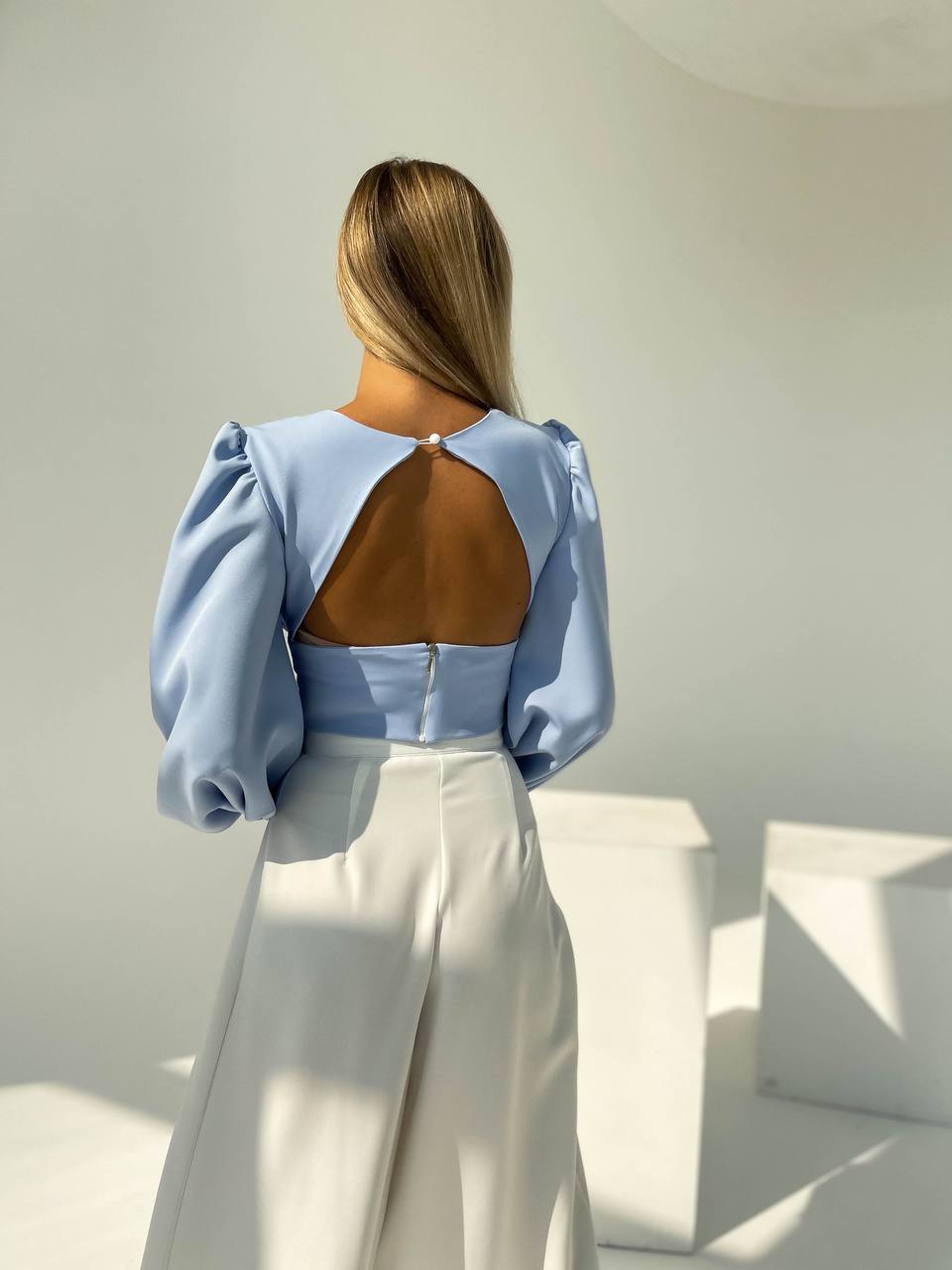 Person wearing a light blue blouse with open back and white pants in a minimal indoor setting