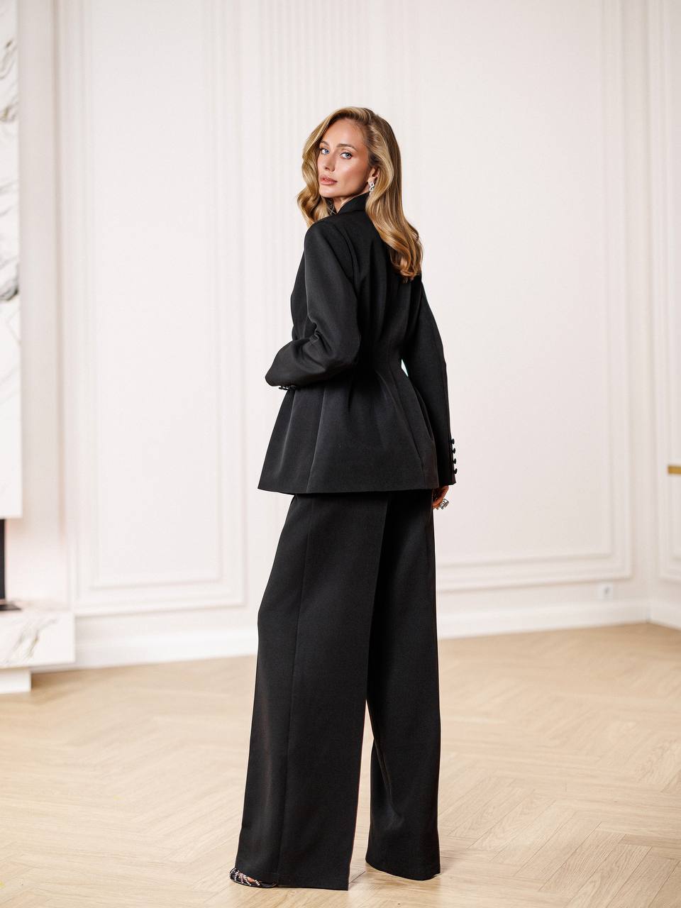 Woman wearing a black suit in an elegant room with white walls and wooden floor.