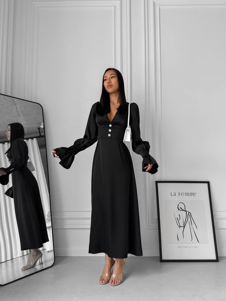 Woman wearing a black dress standing in front of a mirror in a minimalistic room.