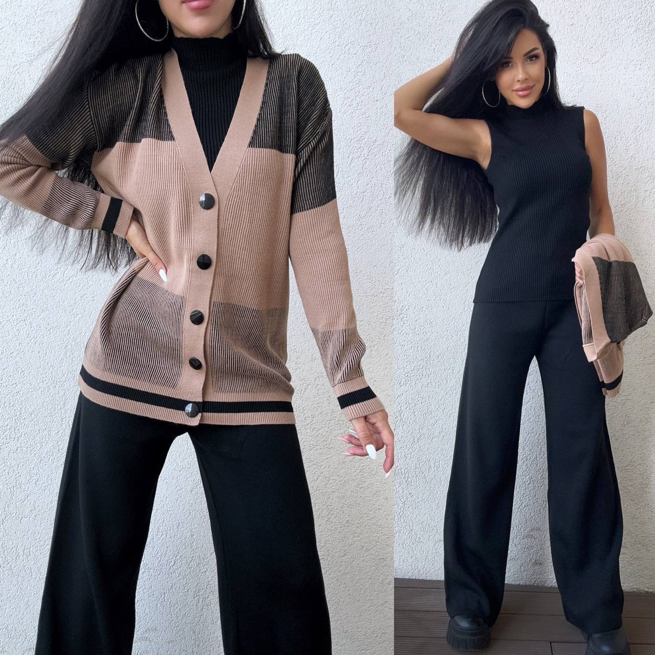 Woman wearing a beige cardigan over a black top with black pants, posing against a light gray wall.