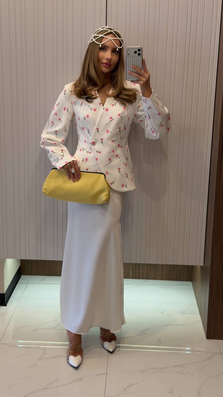 Woman taking a mirror selfie wearing a white floral blouse and white skirt, holding a yellow clutch.