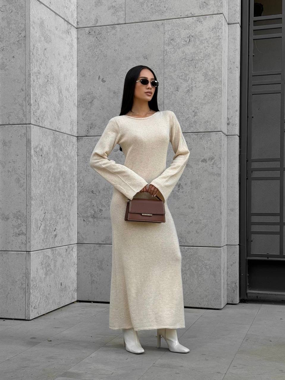 Woman in a long beige dress holding a brown handbag against a gray wall.