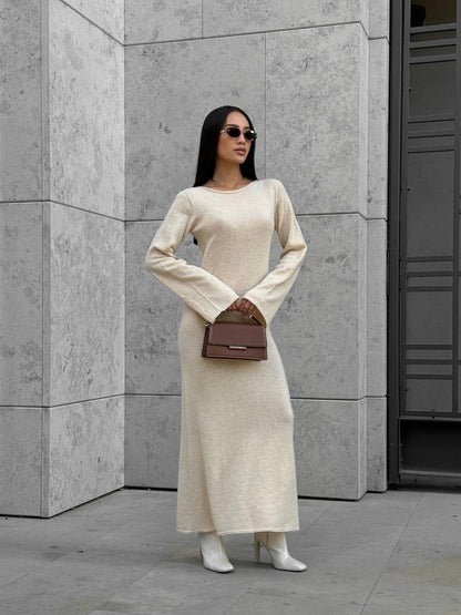 Woman in a long beige dress holding a brown handbag against a gray wall.