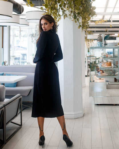 Woman in a black dress standing in a modern indoor setting with white walls and gray furniture.