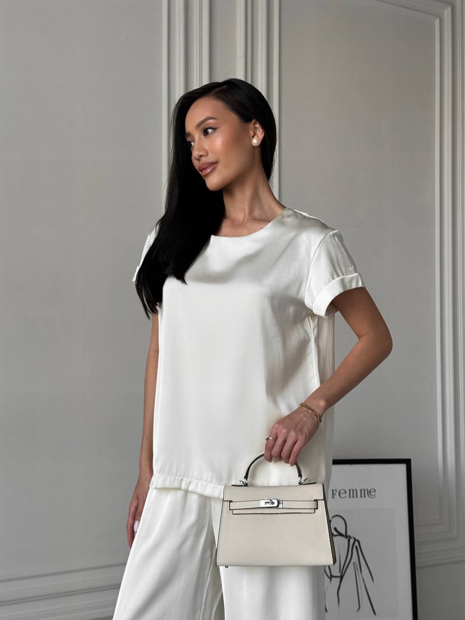 Woman in a white outfit holding a beige handbag against a neutral background