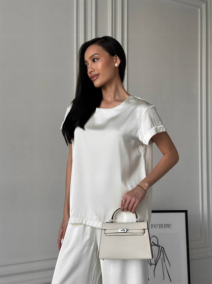 Woman in a white outfit holding a beige handbag against a neutral background