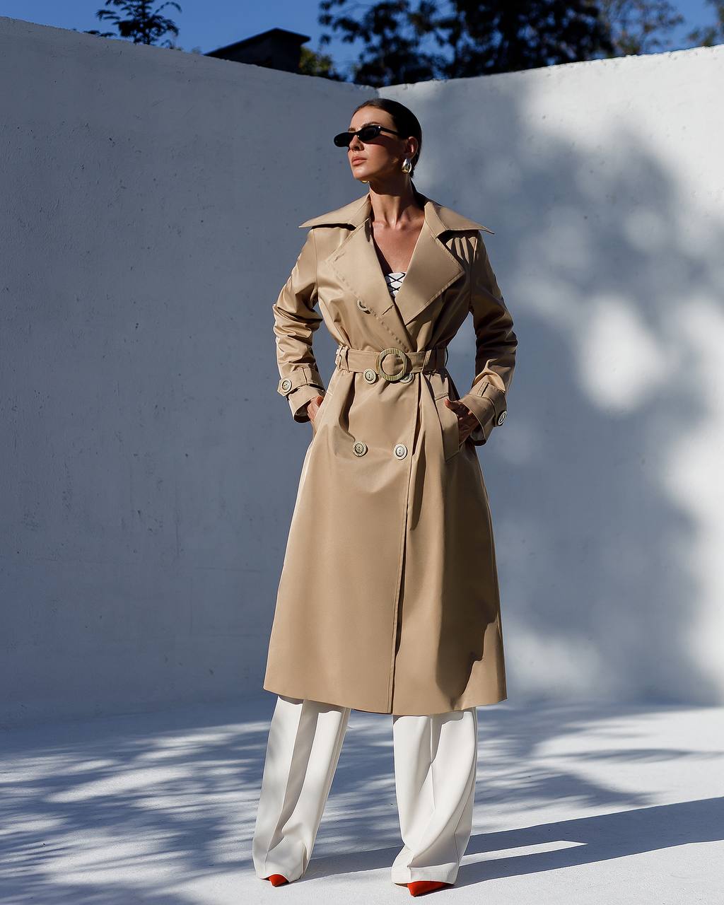 Woman wearing a beige trench coat and white pants against a white wall.
