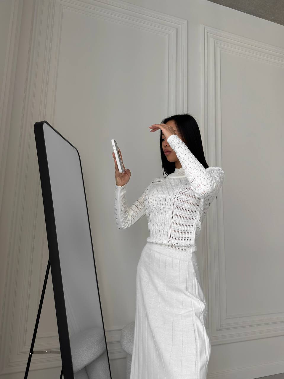 Person taking a mirror selfie wearing a white outfit in a room with decorative wall panels.
