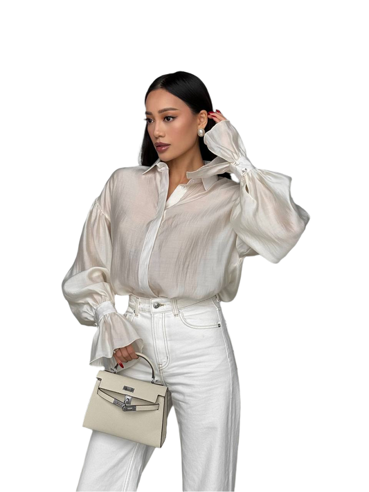 Woman wearing a sheer white blouse and white pants, holding a beige handbag on a white background
