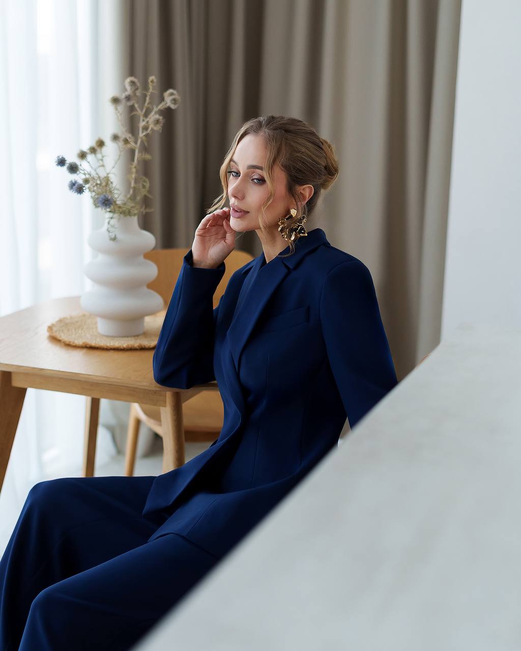 Woman in a navy blue coat sitting indoors with a neutral background