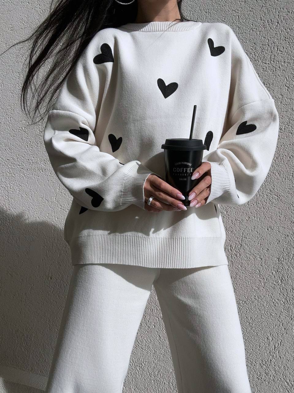 Person wearing a white sweater with black heart patterns, holding a coffee cup against a plain background.