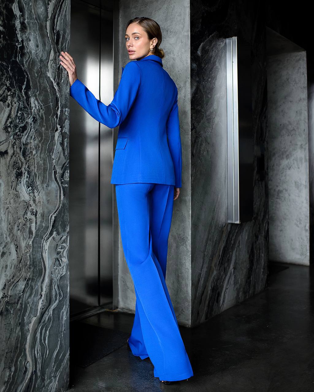 Woman in a blue suit standing against a dark, textured wall.
