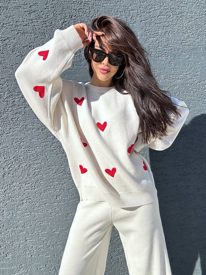 Person wearing a white sweater with red heart patterns against a gray wall.