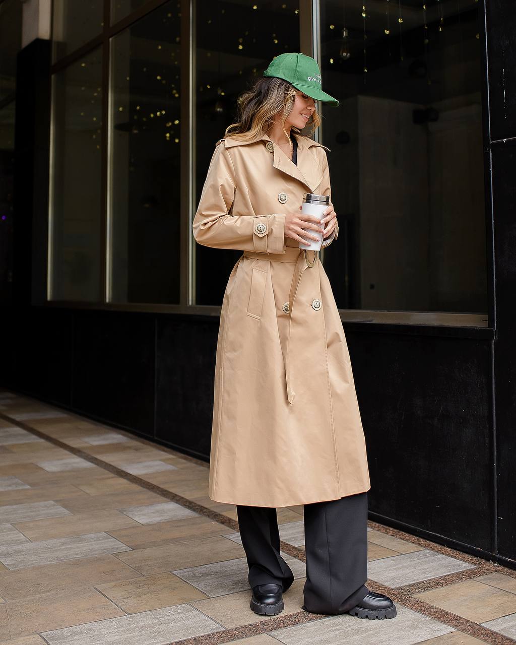 Person wearing a beige trench coat and green cap, holding a coffee cup, standing on a sidewalk.