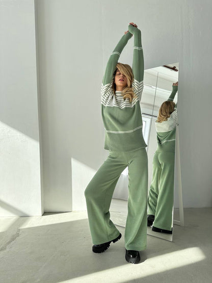 Woman in a green outfit stretching in front of a mirror in a bright room.