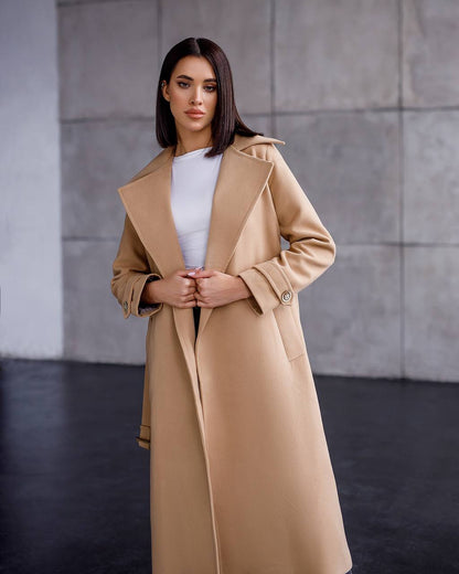 Woman wearing a beige coat against a gray wall.