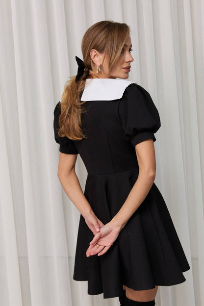 Woman wearing a black dress with a white collar against a white curtain background