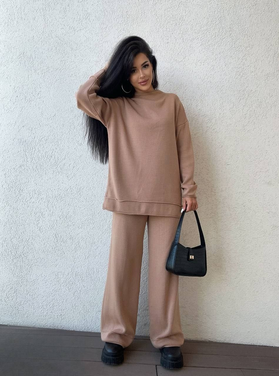 Woman wearing a beige outfit holding a black handbag against a white wall.