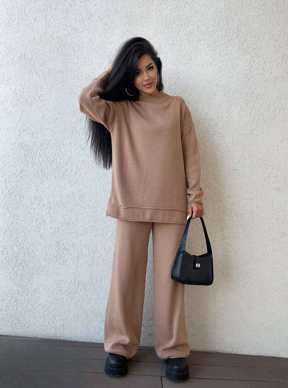 Woman wearing a beige outfit holding a black handbag against a white wall.