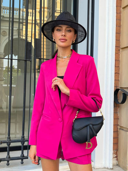 Woman in a bright pink suit with a black hat and handbag, standing in front of a building.