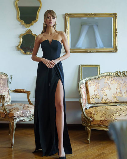 Woman in a black strapless gown with a high slit standing in an elegant room with gold-framed mirrors and a patterned sofa.