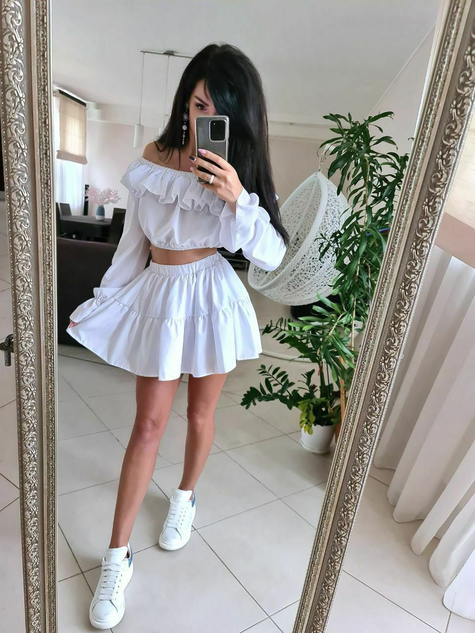 Person taking a mirror selfie wearing a white outfit with a skirt and sneakers in a room.
