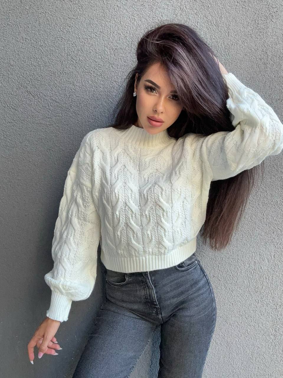 Woman wearing a white cable knit sweater and dark jeans against a gray wall.