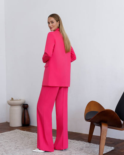 Woman in a bright pink suit standing in a room with a wooden chair and decorative items.