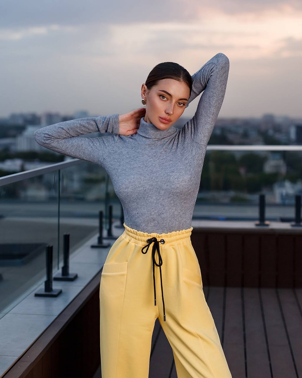 Woman in gray turtleneck and yellow pants on a rooftop with cityscape background
