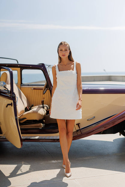 Woman in a white dress standing next to an open vintage car with a clear sky background