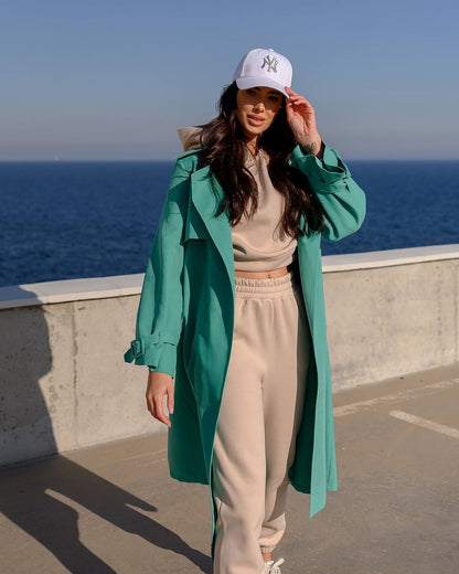 Person wearing a green coat and beige pants with a white cap, standing by the ocean.
