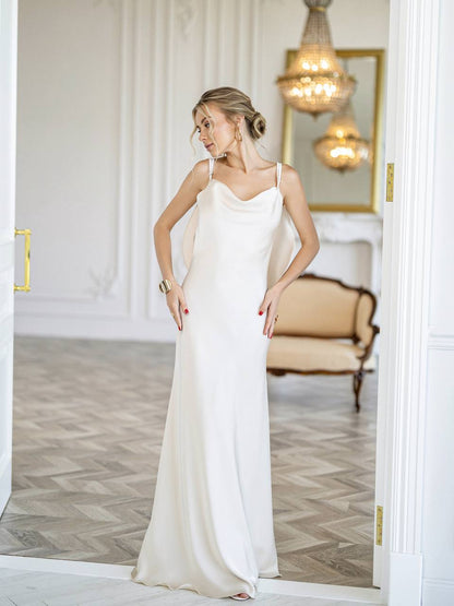 A woman modeling a sleeveless, floor-length white gown with a draped neckline in an elegant indoor room with chandeliers, a mirror, and a bench.