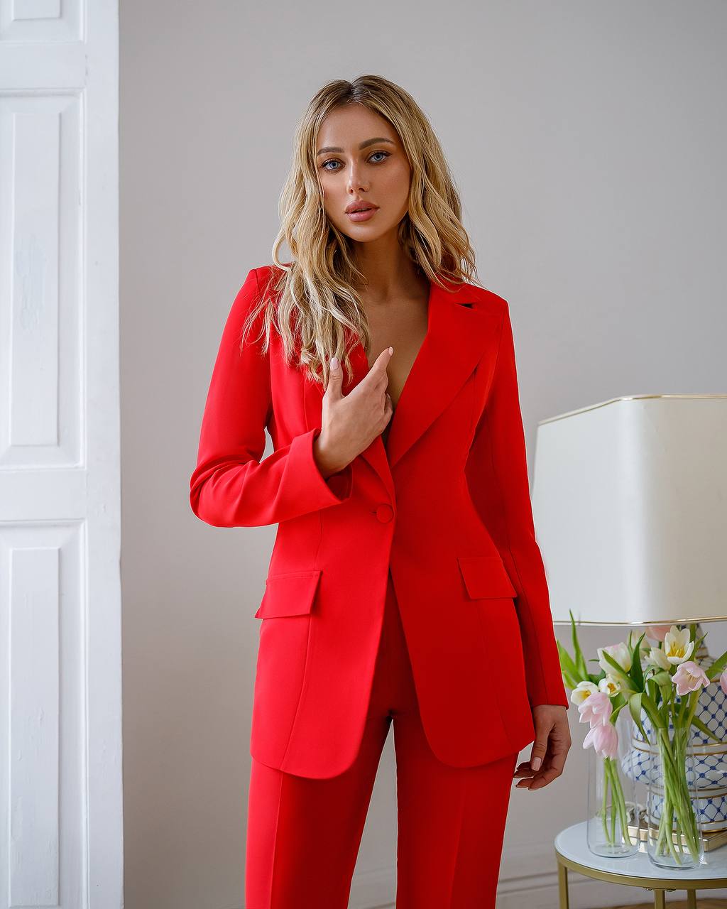 Woman wearing a red suit in an indoor setting with a lamp and flowers in the background.