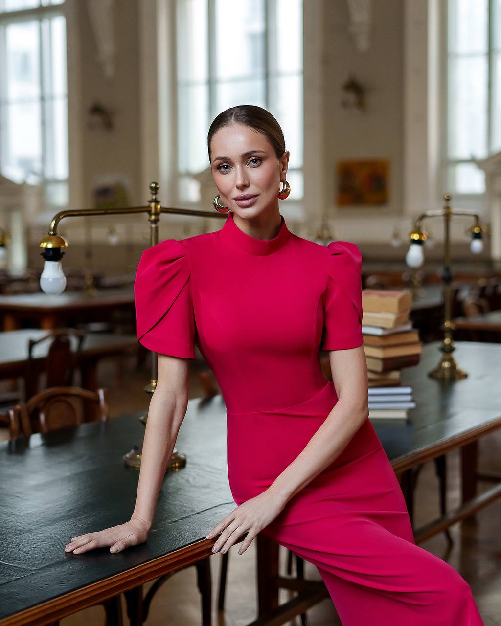 Woman in a bright pink dress standing in an indoor setting with large windows and tables.