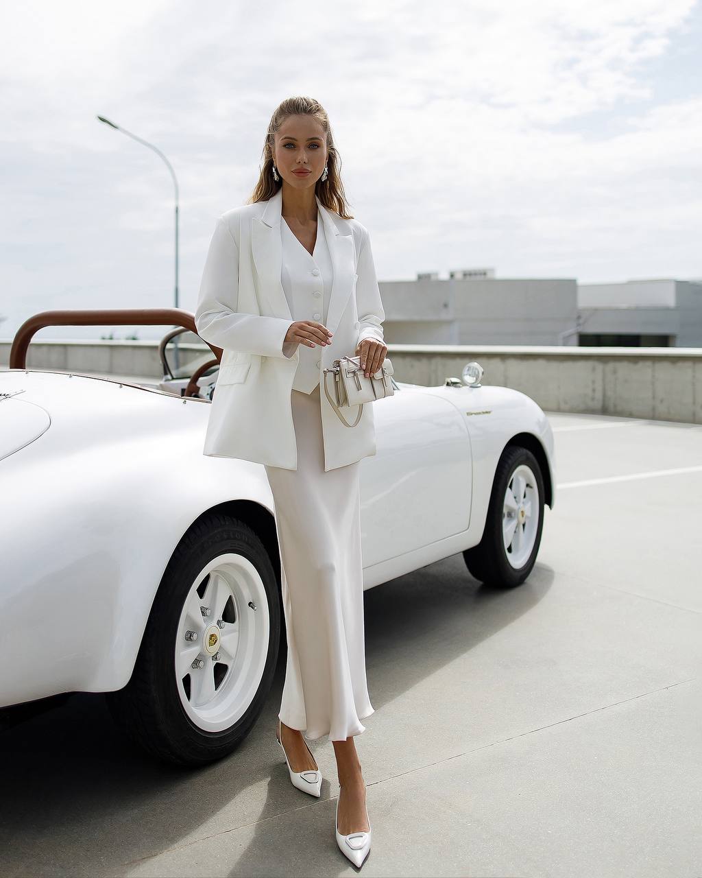 Woman in a white suit standing next to a white car on a rooftop.